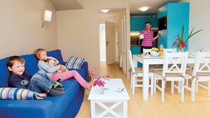 A living room with a blue sofa, a dining table with chairs, and a kitchen area with teal cabinets. - Appartement charmant et sympa | Accès piscine (Cabourg)