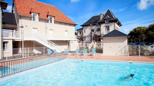 A swimming pool with lounge chairs, a staircase, and a building with a red roof and white walls. - Appartement charmant et sympa | Accès piscine (Cabourg)