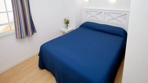 A bedroom with a bed, a small bedside table with a plant, and a window with curtains. - Appartement charmant et sympa | Accès piscine (Cabourg)