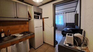 Een compacte keuken met een koelkast, magnetron en spoelbak. Door een open deur is een klein zithoekje met een tafel en stoelen zichtbaar.
