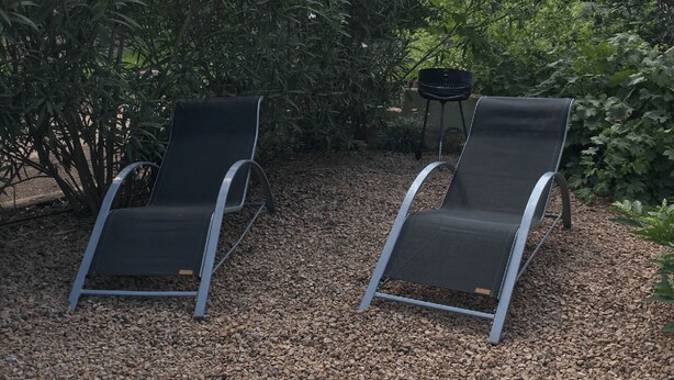 Twee zwarte loungestoelen met metalen frame, geplaatst op een grindpad met weelderige beplanting op de achtergrond.