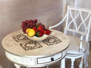 Une table ronde surmontée d’un bol décoratif rempli de fruits, une chaise avec un siège rembourré blanc et un mur en arrière-plan.