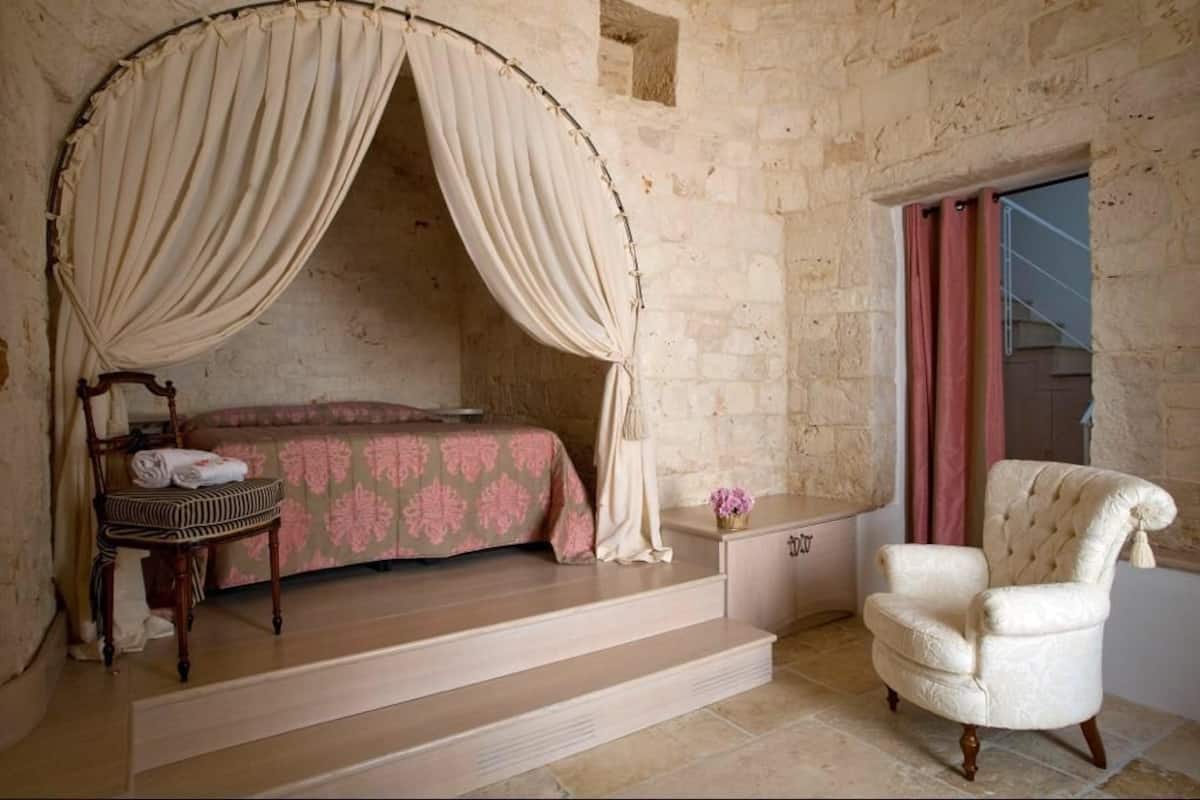 Une chambre à coucher avec un lit à baldaquin, une chaise, une petite table sur laquelle est posé un vase, et un escalier menant à un balcon.