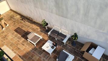 Un patio al aire libre con un sofá, una mesa y una silla, todos de color blanco. Hay dos plantas en macetas y una caja de cartón en el suelo.