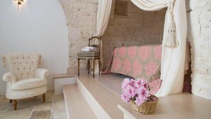 A cozy bedroom with a stone wall, a tufted armchair, a small table, and a bed with a pink patterned bedspread. - Trullo Il Cantastorie (Alberobello)