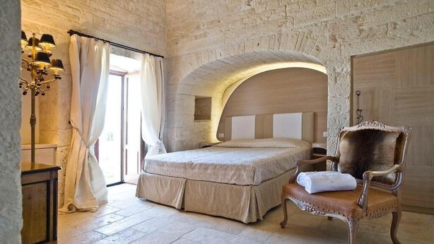 A bedroom with a stone wall, a bed, a chair, and a lamp. - Trullo Il Cantastorie (Alberobello)