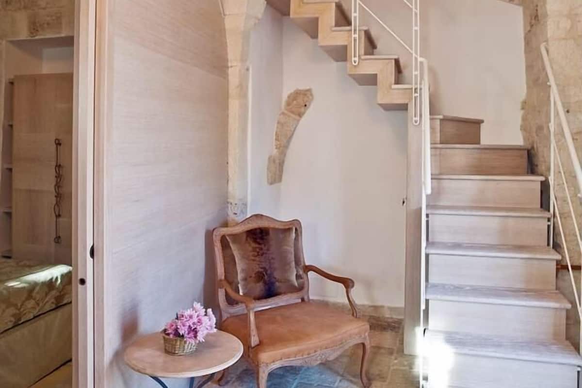 Une pièce aux murs de pierre, avec une chaise en bois et une petite table, un escalier doté d’une rampe en métal, et une porte donnant sur une autre pièce.