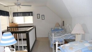 Ein kleiner, gut beleuchteter Raum mit einem Einzelbett, einem Schreibtisch, einem Stuhl, einer Lampe und einem Deckenventilator.