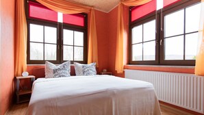 Ein Schlafzimmer mit einem großen Bett, zwei mit Vorhängen versehenen Fenstern, einem Nachttisch und einem Heizkörper.