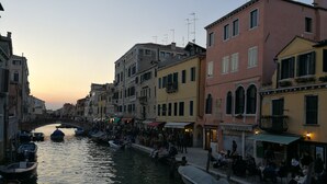 A canal with boats docked, a bridge, and buildings with shops and outdoor seating. - Casa Elia Venice ... (Venezia)