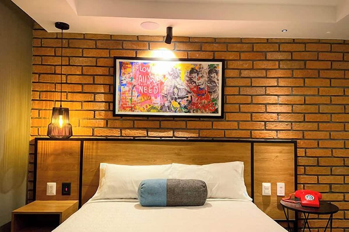 A hotel room with a brick-patterned wall, a bed with white and grey bedding, a colorful artwork above the bed, a small table with a red telephone, and a wall-mounted light fixture.