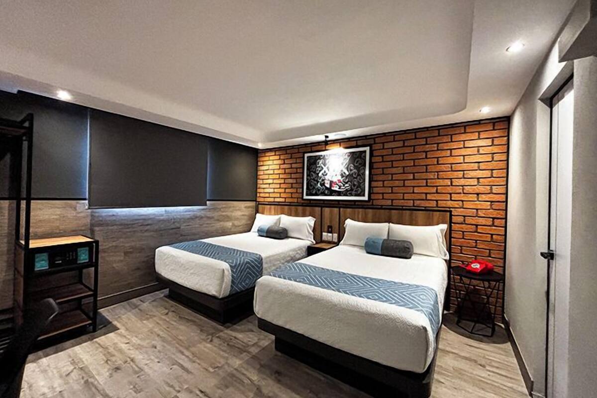 A modern hotel room with two beds, a brick accent wall, a mounted TV, and a small table with a red object.