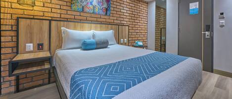 A modern hotel room with a brick-patterned wall, a large bed with patterned bedding, a bedside table, a nightstand, a wall-mounted lamp, and a door with a lock.