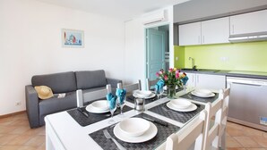 A dining area with a table set for four, a grey sofa, a kitchen with white cabinets, and a green wall. - Accès Piscines ! Appartement cosy et charmant (Grimaud)