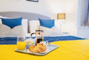 Bandeja de desayuno con cruasanes, una cafetera y un vaso de jugo de naranja sobre una cama con colcha azul y amarilla.