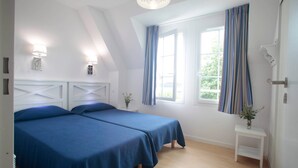 A room with two beds, a window with curtains, a small table with a plant, and a white headboard. - Appartement cozy, près de la plage (Cabourg)