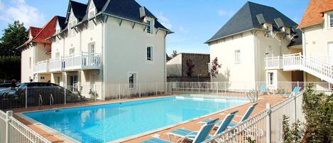 A swimming pool with lounge chairs, surrounded by white fences and buildings with dark roofs.