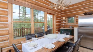 A log cabin with a dining area featuring a large table set for four, a refrigerator, and a view of the forest through large windows. - Red Cloud 4 | Ski-in/Out Cabin, Hot Tub, Sleeps 12 (Big Sky)