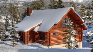 A log cabin with a snow-covered roof, surrounded by snow-covered trees and mountains in the background. - Red Cloud 4 | Ski-in/Out Cabin, Hot Tub, Sleeps 12 (Big Sky)