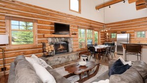 A log cabin interior with a stone fireplace, wooden walls, a dining area, and a kitchen. - Red Cloud 4 | Ski-in/Out Cabin, Hot Tub, Sleeps 12 (Big Sky)