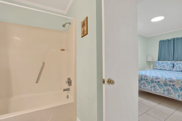 A bathroom with a bathtub and shower, a bed with a floral bedspread, and a small framed picture on the wall. - Unit 217b, 1 bedroom 1 bath condo, Bay/Beach front condo (Tybee Island)