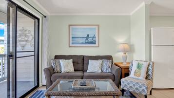 A living room with a sofa, armchair, and coffee table. There is a framed picture on the wall, a lamp, and a refrigerator.