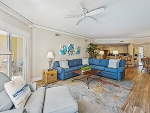 A living room with a blue sectional sofa, a coffee table, and a floor lamp. There is a ceiling fan, a dining area with chairs, and a kitchen in the background.