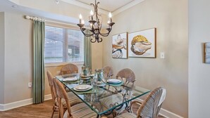 A dining room with a glass table, wicker chairs, a chandelier, and artwork on the walls.
