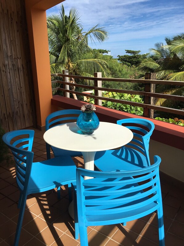 Et rundt bord med en blå vase, omgivet af blå stole, på en terrasse med udsigt til palmer og et træhegn.