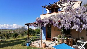 Une villa dotée d’une pergola recouverte de fleurs violettes, d’une terrasse avec une table et des chaises, et offrant une vue sur la campagne.