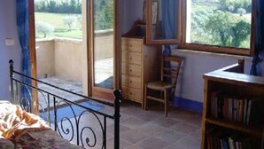 Une chambre à coucher comprenant un lit, une commode, une chaise et une bibliothèque. Il y a deux fenêtres avec des rideaux, et on aperçoit un balcon à travers l’une des fenêtres.