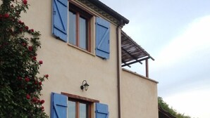 Une maison à deux étages avec des volets bleus, une grande plante en pot au premier plan et un patio couvert.