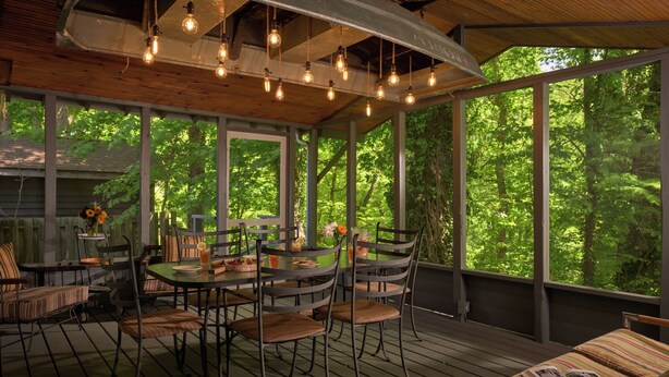 A covered outdoor dining area with a table and chairs, surrounded by large windows offering a view of a forested area. - River's Edge on 30 Acres with Kayaks/Pool/Farm/River/Trails/Pets, near Beach (Union Pier)