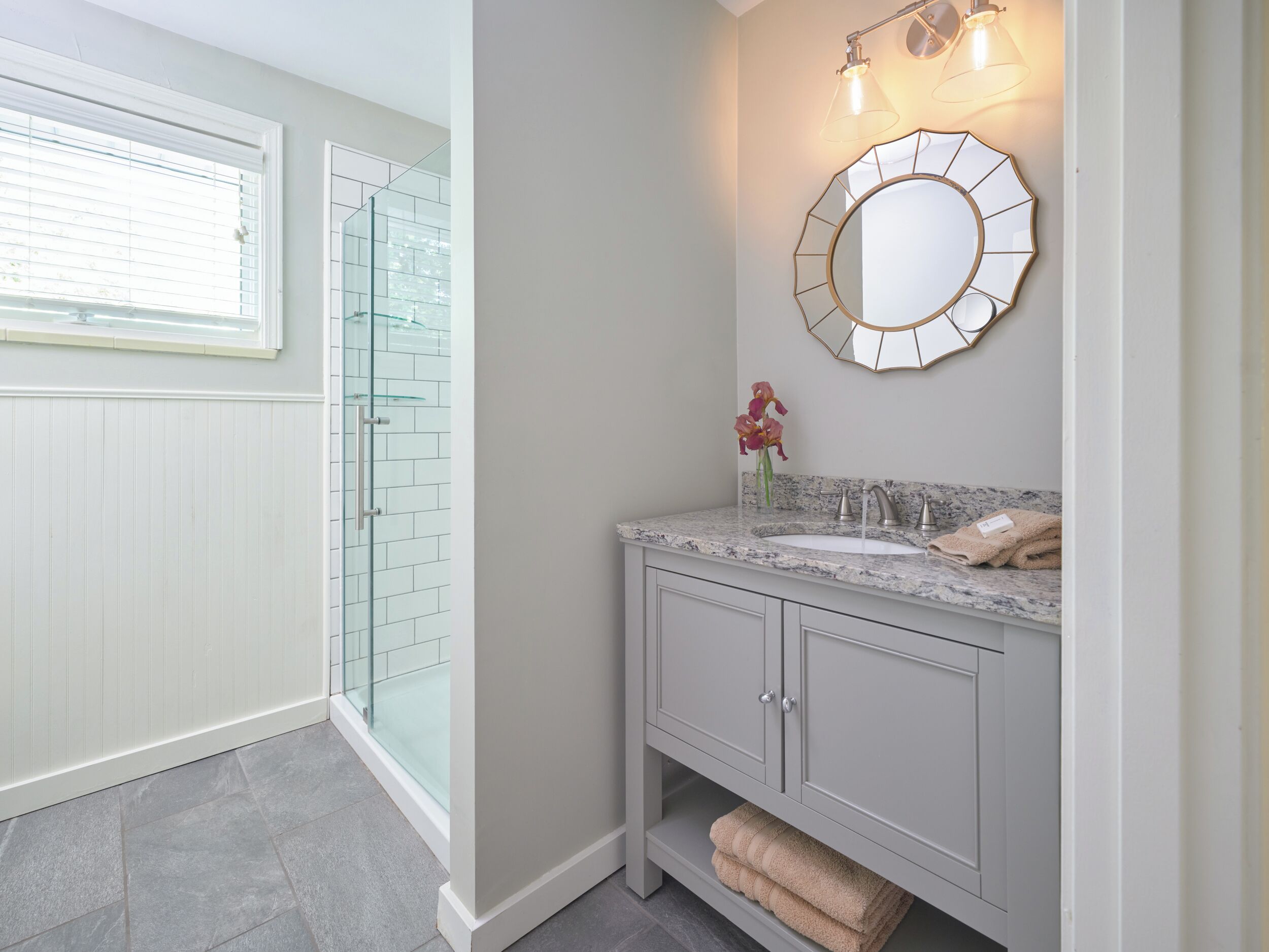 A bathroom with a glass shower enclosure, a vanity with a marble countertop, and a wall-mounted mirror.