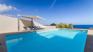 Ein klarer, blauer Pool mit Liegestühlen und Sonnensegel, mit Blick auf den Ozean.