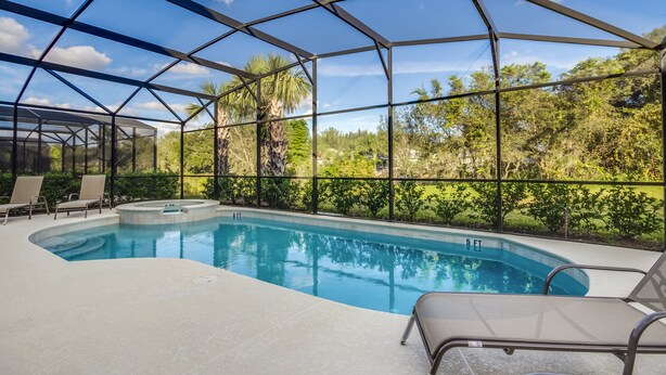 A covered outdoor swimming pool with clear blue water, surrounded by a metal frame and glass panels, with lounge chairs and a hot tub.