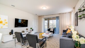 A modern living room with a flat-screen TV, a dining area with a white table and black chairs, and a grey sofa with a white orchid plant. - Solterra Exec Villas- 5445misty (Davenport)