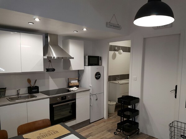 A compact kitchen with white cabinets, a stainless steel range hood, a microwave, and a refrigerator. There's a small dining area with a table and chairs. - VACATION HOME, EVERYTHING BY BIKE! (NOIRMOUTIER)