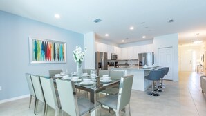 Una cocina moderna con gabinetes blancos, electrodomésticos de acero inoxidable y un comedor con mesa y sillas de vidrio.