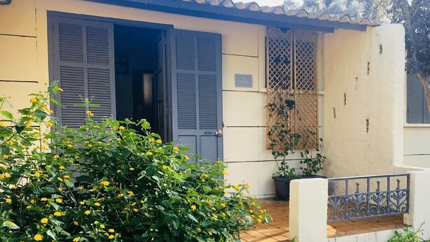 Un edificio con dos puertas abiertas, un pequeño balcón con barandilla de metal y una planta en maceta a la derecha.