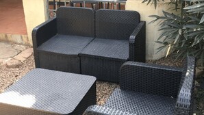 Un sofá de mimbre negro y un reposapiés a juego en un patio exterior con suelo de concreto y baranda metálica.