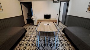 Una habitación pequeña con una mesa de madera, dos sofás grises, un televisor y un ventilador.