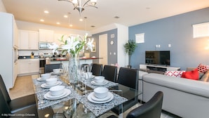 Une cuisine moderne avec des meubles blancs, un coin repas doté d’une table en verre pour six personnes, et un salon avec un canapé et un téléviseur.