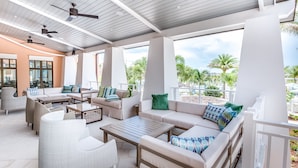 Un espace de détente extérieur doté de ventilateurs de plafond, de sièges confortables et de grandes fenêtres offrant une vue sur les palmiers et la plage.