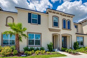 A two-story house with a beige exterior, multiple windows, and a well-maintained front yard with various plants and a palm tree. - Solara Resort Exec Villas- 1542cpc (Kissimmee)
