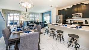 A modern kitchen with dark cabinets, a microwave, and a refrigerator. There is a dining area with a table set for four and four bar stools. - Champions Gate Exec Villas- 8917stinger (Davenport)
