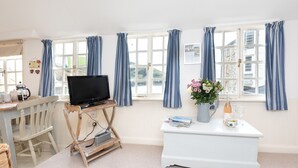 A room with a television on a wooden stand, a white table with a vase of flowers, and a window with blue curtains. - Harbour Loft - Two Bedroom Apartment, Sleeps 4 (Port Isaac)