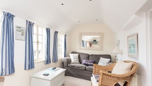 A cozy living room with a grey sofa, a white side table, and a wicker chair. There are blue curtains, a mirror, and a framed picture on the wall. - Harbour Loft - Two Bedroom Apartment, Sleeps 4 (Port Isaac)