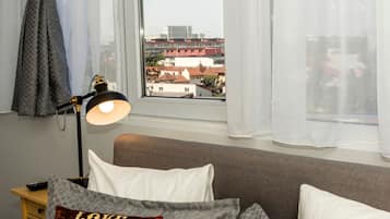 Ein Schlafzimmer mit Blick auf Gebäude durch ein Fenster, eine Lampe auf einem Nachttisch und ein Kissen mit Schrift.