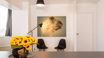 Ein modernes Zimmer mit weißer Wand, einem großen gerahmten Kunstwerk, zwei schwarzen Stühlen, einem Tisch mit einer Vase voller Sonnenblumen und einer Pendelleuchte.